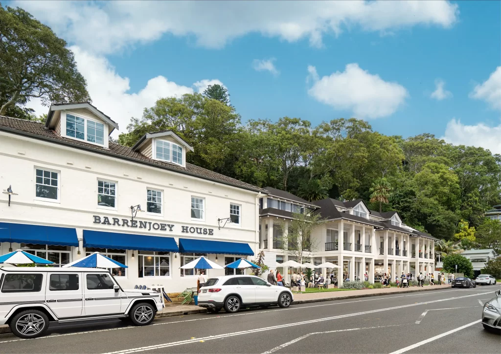 Street view of Barrenjoey House and adjacent buildings in Palm Beach, surrounded by trees with cars parked along the roadside and people enjoying the outdoor seating.