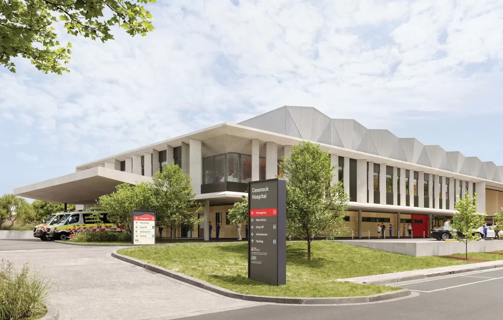 Rendering of the proposed Cessnock Hospital redevelopment featuring a two-storey modern building with geometric roof design, landscaped greenery, ambulance parking bays, and a clear signage system at the main Jurd Street entrance.