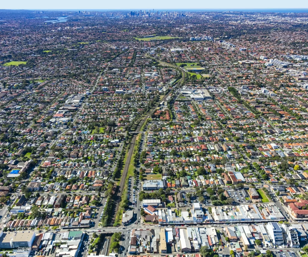 Aerial view of the Canterbury-Bankstown area in Sydney, showing a mix of residential homes, commercial buildings, green spaces, and transport corridors with the Sydney city skyline visible in the background.