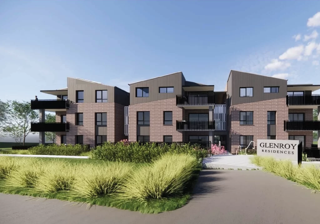 A three-story modern residential building with brick and metal cladding exterior, featuring multiple balconies and surrounded by lush landscaping. A sign in the foreground reads "Glenroy Residences."