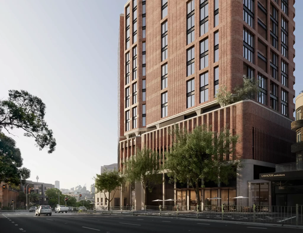 A modern 17-storey mixed-use building with a brick façade and large windows, located on a busy street corner. The lower levels feature retail spaces with outdoor seating, and greenery is integrated into the design. Cars are seen on the road, with the Sydney skyline, including the Sydney Tower, visible in the background.