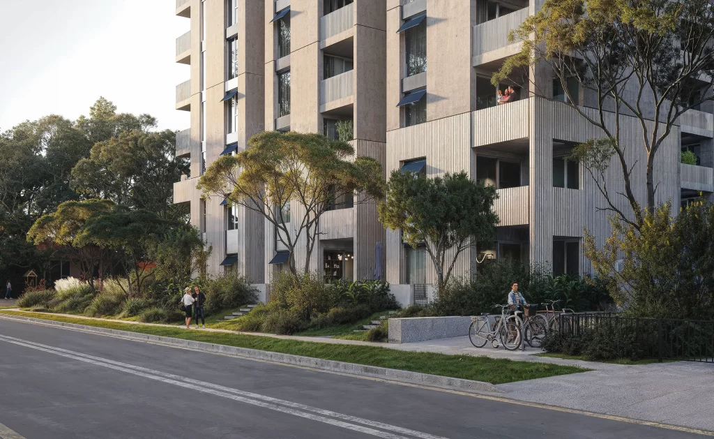 A modern mid-rise apartment building with a textured concrete facade, large balconies, and integrated greenery. The building is surrounded by landscaped gardens with mature trees and native plants. A pedestrian walkway with a bike rack is in the foreground, where a person is parking their bicycle. Residents can be seen on balconies, and two people are conversing near the entrance. The setting appears to be in a suburban or urban residential area with a strong focus on greenery and sustainability.