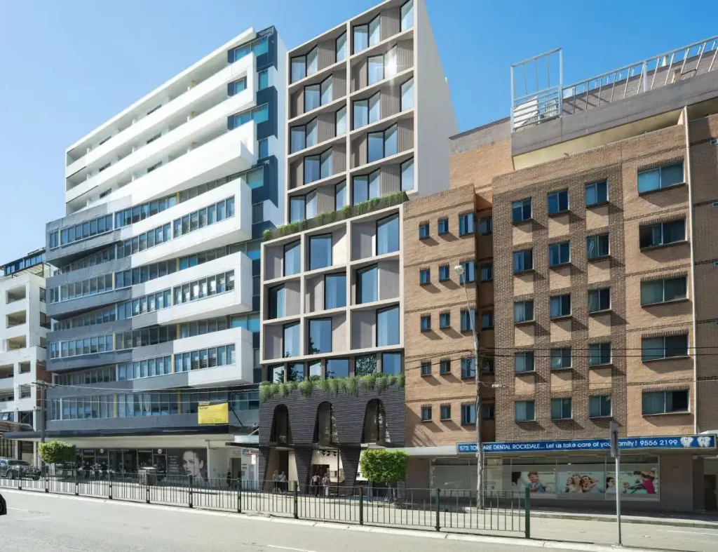 A rendered image of a proposed 12-storey mixed-use development in Rockdale, NSW, featuring a modern façade with large windows, greenery on terraces, and a ground-floor commercial space. The building is situated between two existing structures, one a contemporary white apartment complex and the other a traditional brown brick residential building.