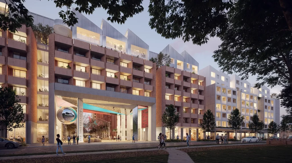 Architectural rendering of The Timberyards development in Marrickville, showing a modern multi-story residential complex with warm-toned brick facades, large balconies, and rooftop terraces. The entrance features a large sculptural archway with integrated public art, leading into a well-lit open courtyard with greenery and communal spaces. Pedestrians and cyclists are seen moving along landscaped pathways, activating the surrounding streetscape.