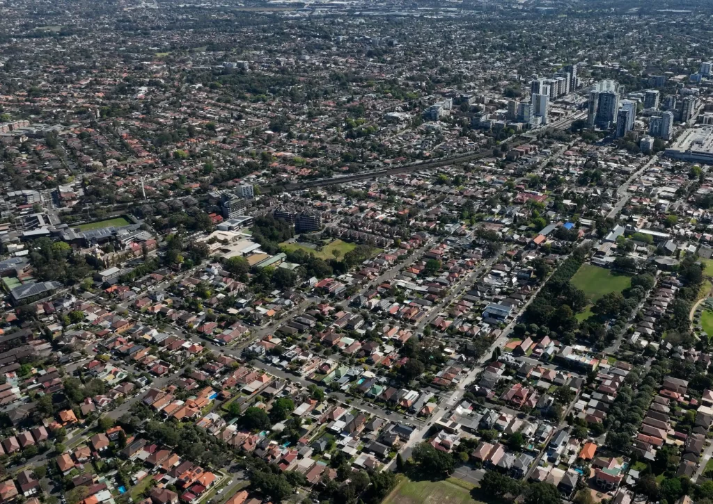 Aerial view of Croydon, Sydney, showcasing a suburban landscape with low-rise residential housing, tree-lined streets, and pockets of green space. In the background, high-rise buildings in Burwood contrast with the predominantly low-density urban fabric.