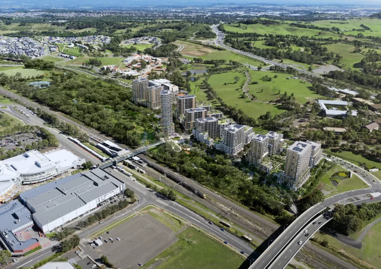 Aerial rendering of the proposed Macarthur Gardens North development in Campbelltown, showing high-rise residential buildings surrounded by green spaces, with Macarthur Train Station, a shopping centre, and road networks in the foreground.