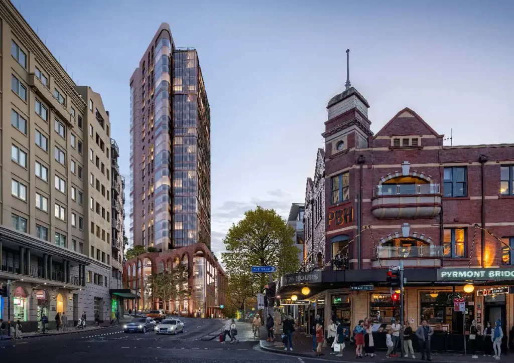 A rendered image of the approved Pyrmont Over Station Development, a modern high-rise tower with a curved glass facade and a brick podium. The tower is set against a heritage streetscape, including the Pyrmont Bridge Hotel, at dusk with pedestrians and cars activating the area.