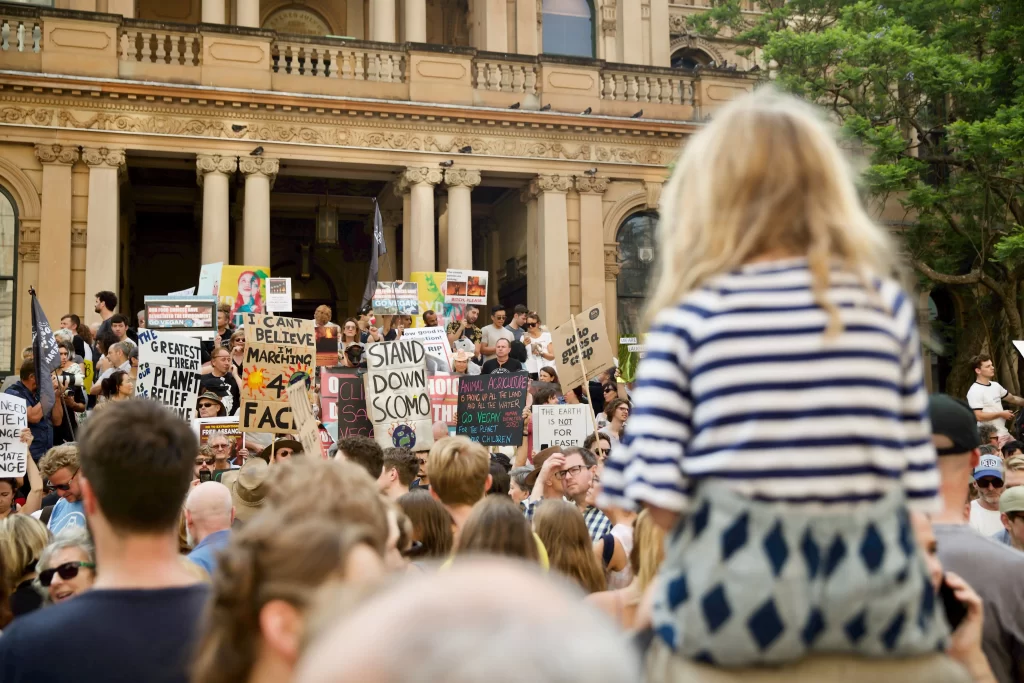 A large crowd gathers in front of a historic sandstone building, holding signs and banners related to climate change and environmental activism. A young child sits on someone’s shoulders, observing the protest. The signs display messages advocating for climate action, environmental protection, and political change.