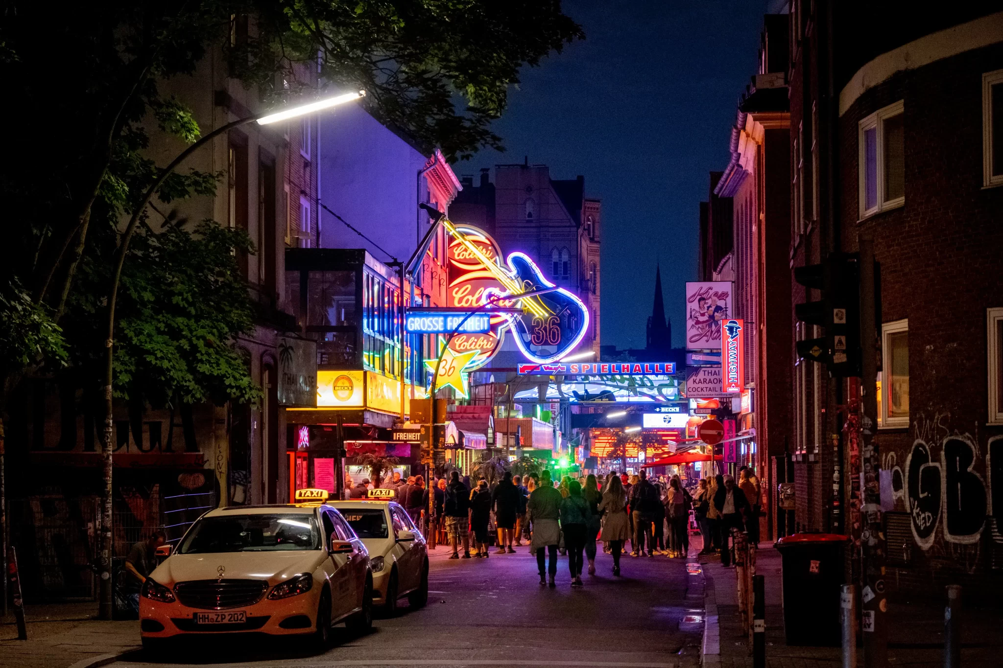Crowds walk along a neon-lit nightlife street with bars, clubs, and taxis at night in an urban entertainment district.